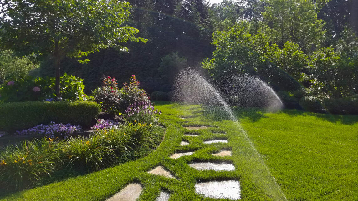 A garden pathway lined with square stepping stones cuts through a lush lawn as a sprinkler system waters the area